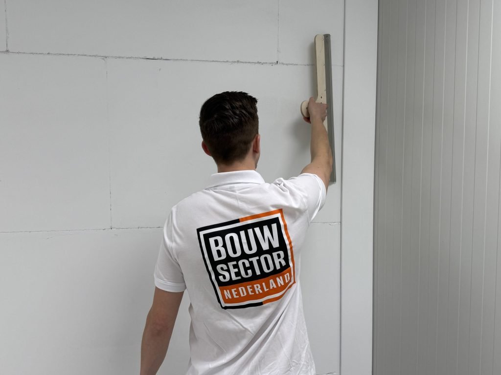 Achteraanzicht van een stukadoor met het logo van Bouwsector Nederland op zijn shirt, die een wand met een lange spaan afreit
