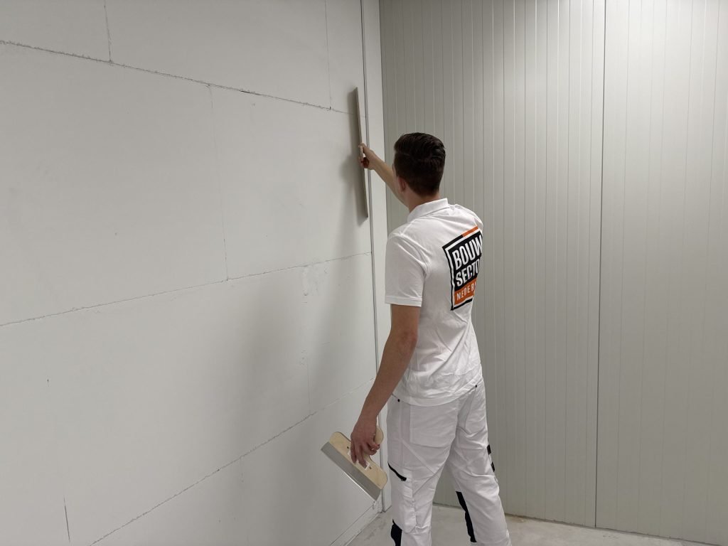 Stukadoor houdt een lange spaan boven zijn hoofd tegen een lichte wand en heeft een brede plamuurspaan in zijn linkerhand
