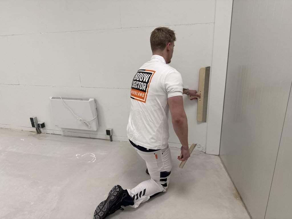Stukadoor zit op een knie en trekt de pleister vlak bij de vloer glad met een brede spaan