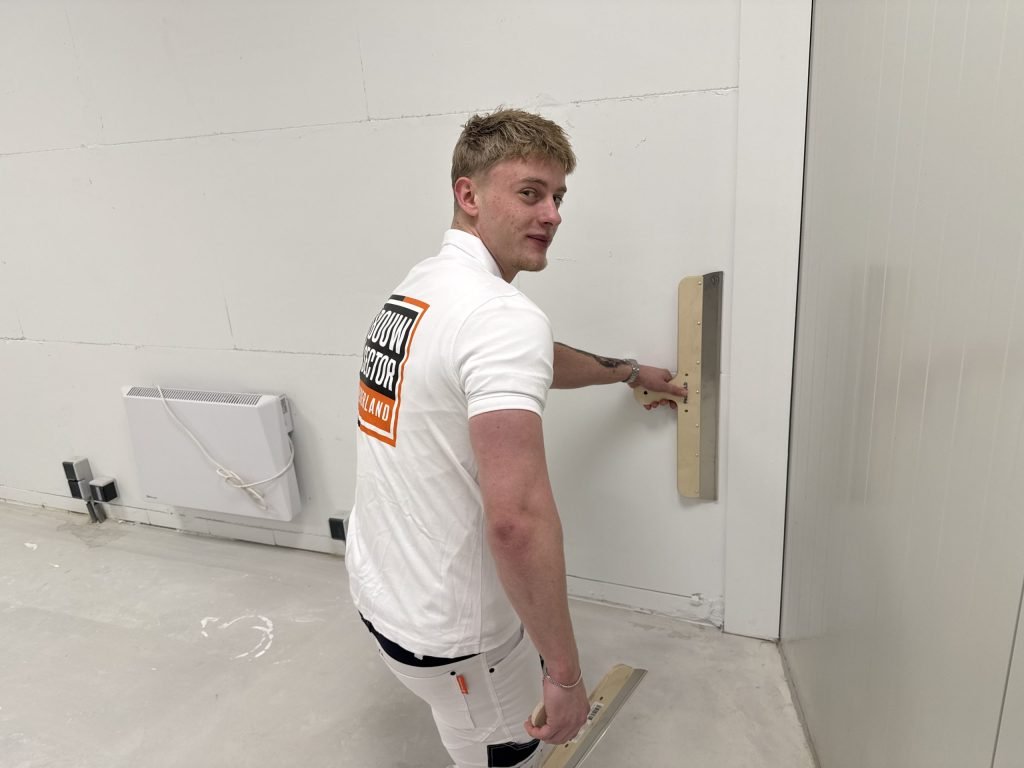 Jonge stukadoor kijkt om naar de camera terwijl hij met een brede spaan een wand vlak bij de hoek afwerkt