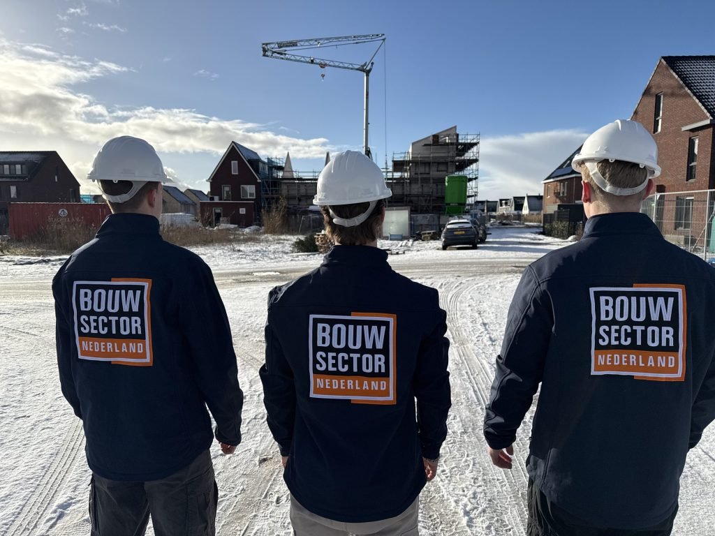 Drie collega's in werkkleding met oranje-wit logo overzien een bouwplaats in aanbouw